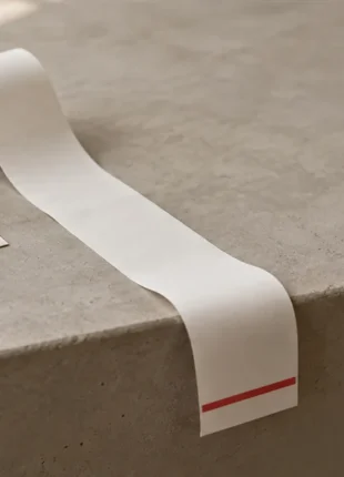 A long blank till receipt unrolling across a pale stone ledge and curling over the front edge, one scarlet band near its end, an empty card lying to the left: spend recorded, Google Ads not converting into enquiries