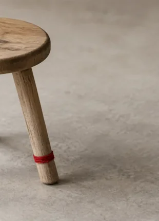 A small plain wooden three-legged stool standing on a warm grey surface with a thin scarlet cord tied around one leg, an illustration of SEO vs AEO vs GEO as three legs under one seat